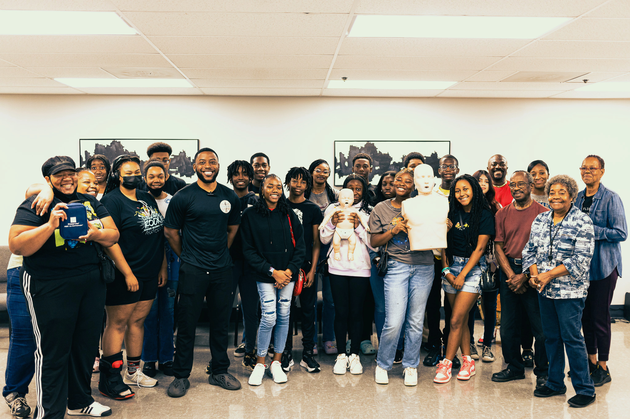 GoodWorks founder poses with a large group of community members after CPR training, with adult and infant manikins