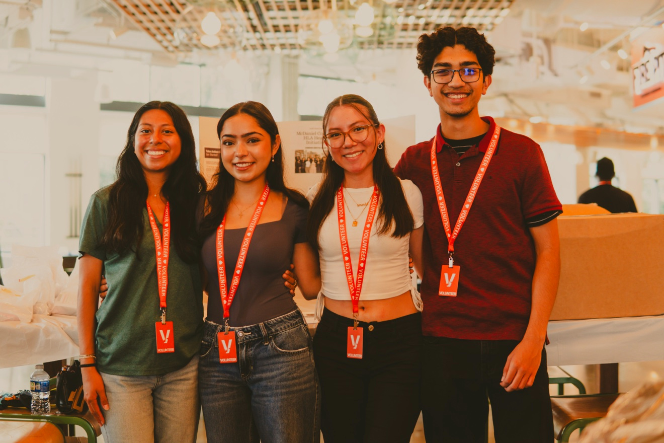 Four smiling GoodWorks volunteers wearing red lanyards stand together at a community event